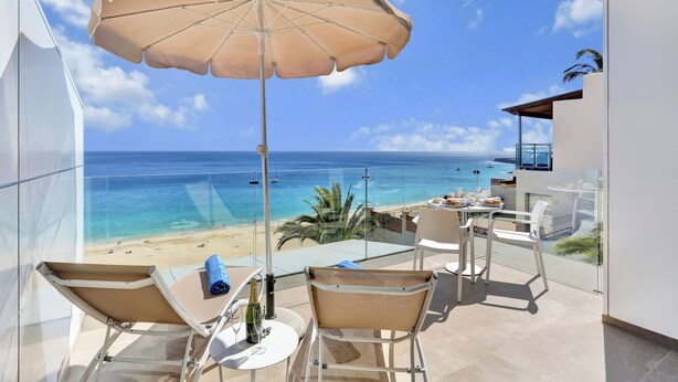 Ein Balkon mit Blick auf Strand und Meer, ein Tisch mit Stühlen und ein großer Sonnenschirm.