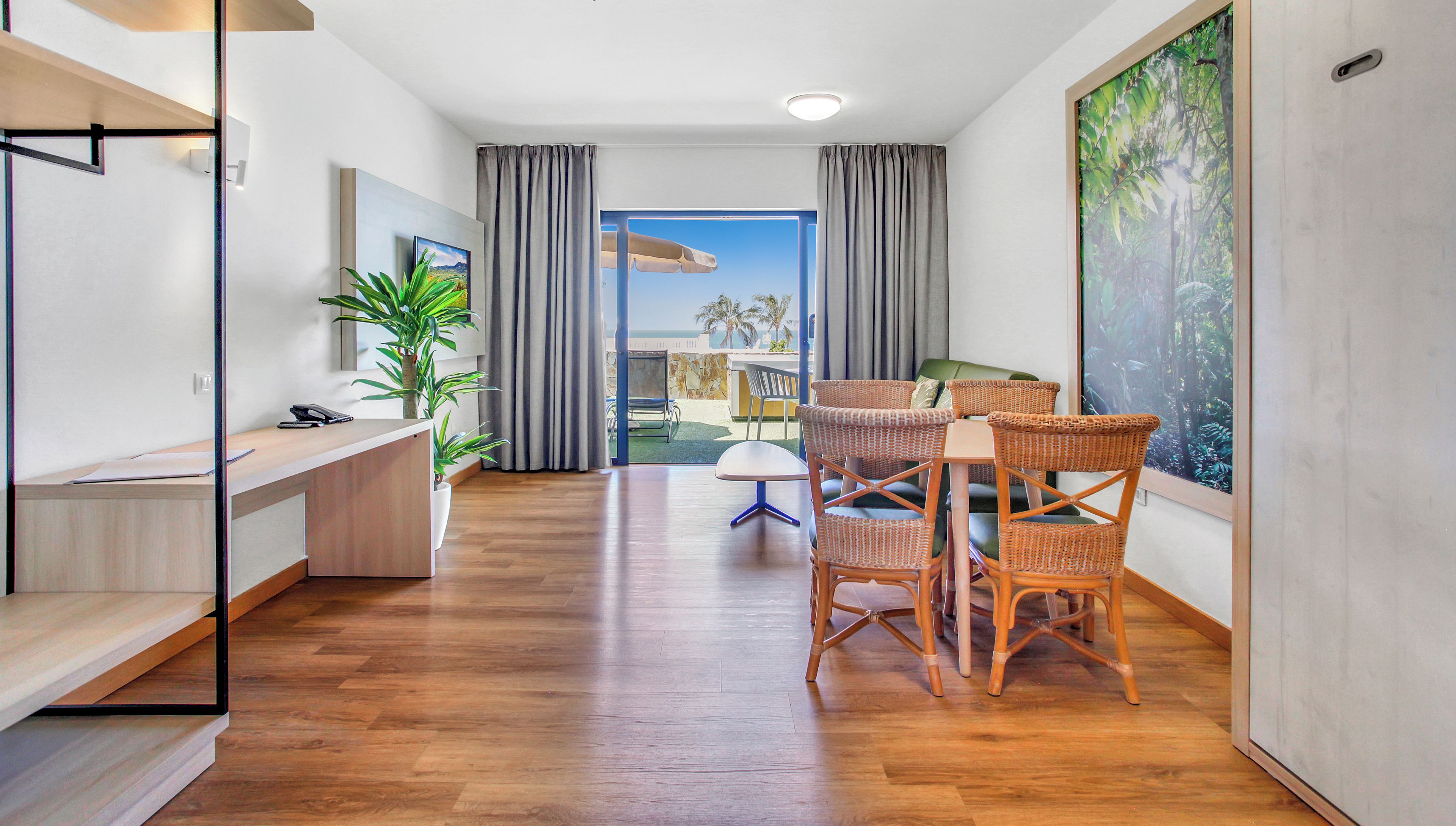 Une chambre d’hôtel moderne avec un plancher en bois, un bureau avec un téléphone et des documents, des fauteuils en osier, une petite table et une grande fenêtre avec des rideaux.