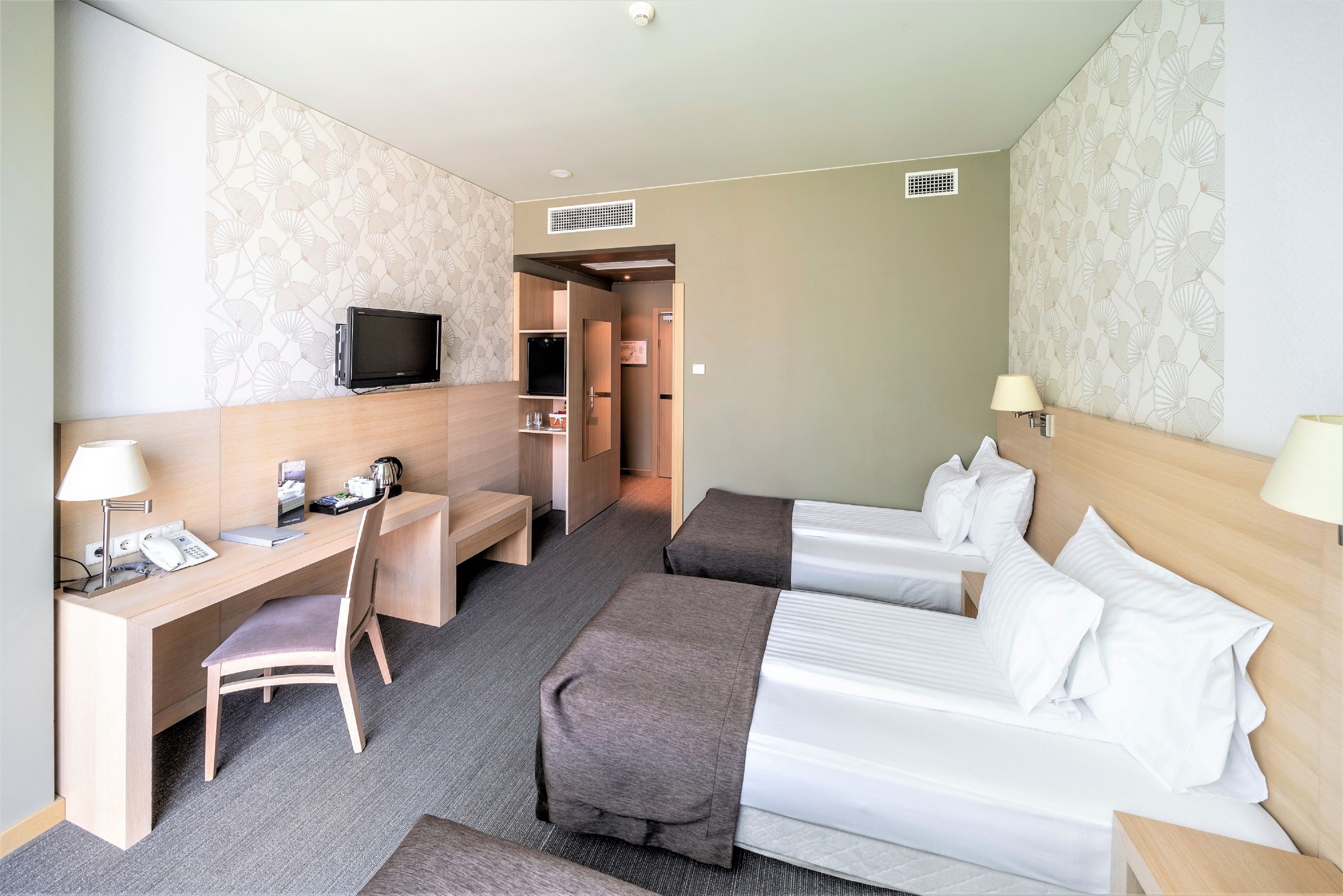 Una camera d'albergo con una scrivania, due letti, una sedia, una TV e una lampada.