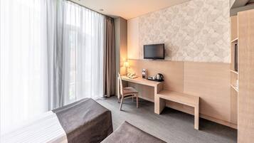 A hotel room with a bed, a desk with a chair, a TV mounted on the wall, and a window with curtains.