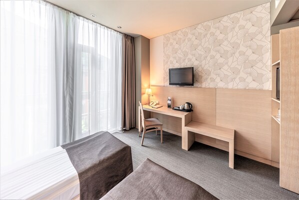 A hotel room with a bed, a desk with a chair, a TV mounted on the wall, and a window with curtains.
