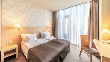 Hotellihuone, jossa on suuri sänky, kaksi yöpöytää valaisimineen, työpöytä tuolilla sekä ikkuna, jossa on verhot.
