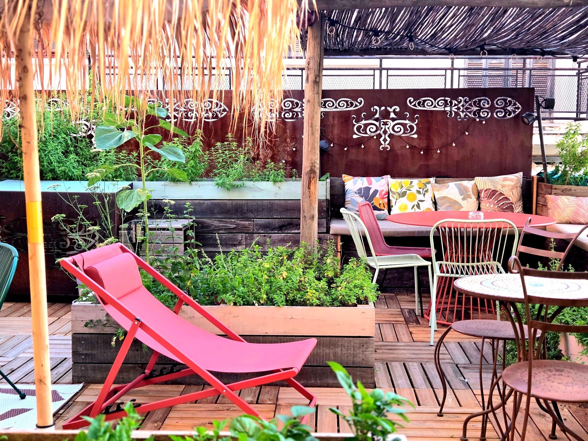 Un espace patio doté d’une terrasse en bois, d’un fauteuil de jardin rouge, d’une table en métal avec des chaises et d’un mur décoratif orné de motifs.