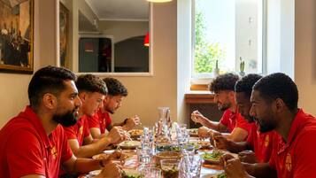 A group of people in red shirts dining at a table with various dishes and glasses, in a room with a large mirror and a window.