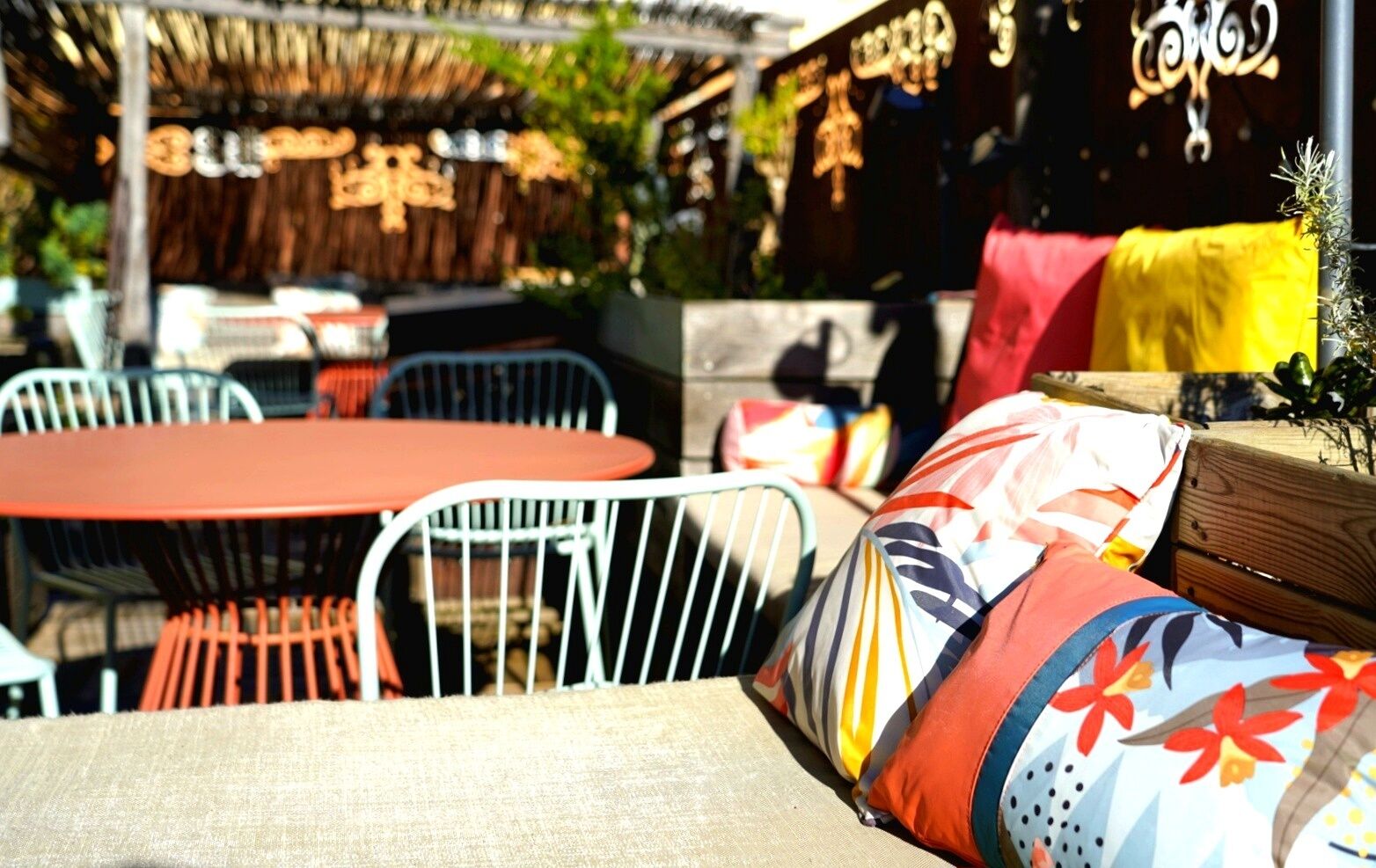 An outdoor seating area with metal chairs and tables, a thatched roof, and a wooden bench with colorful cushions.