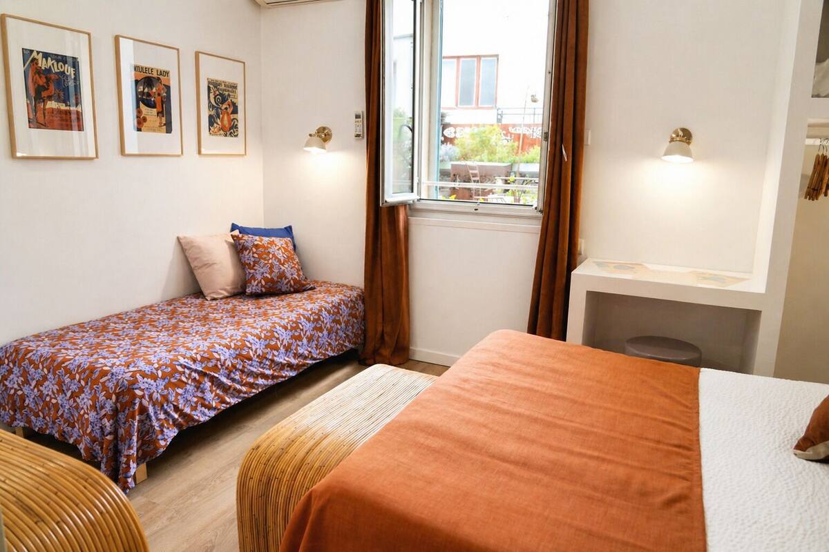 A room with two beds, a window with curtains, wall-mounted lights, and framed posters.