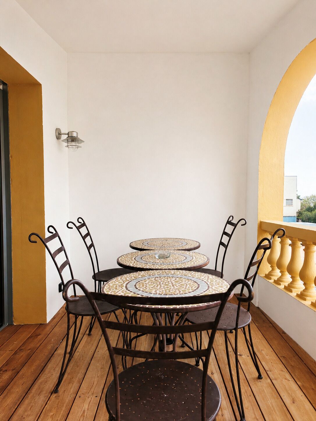 A balcony with a wooden floor, a white wall, a yellow arch, and a table with chairs.