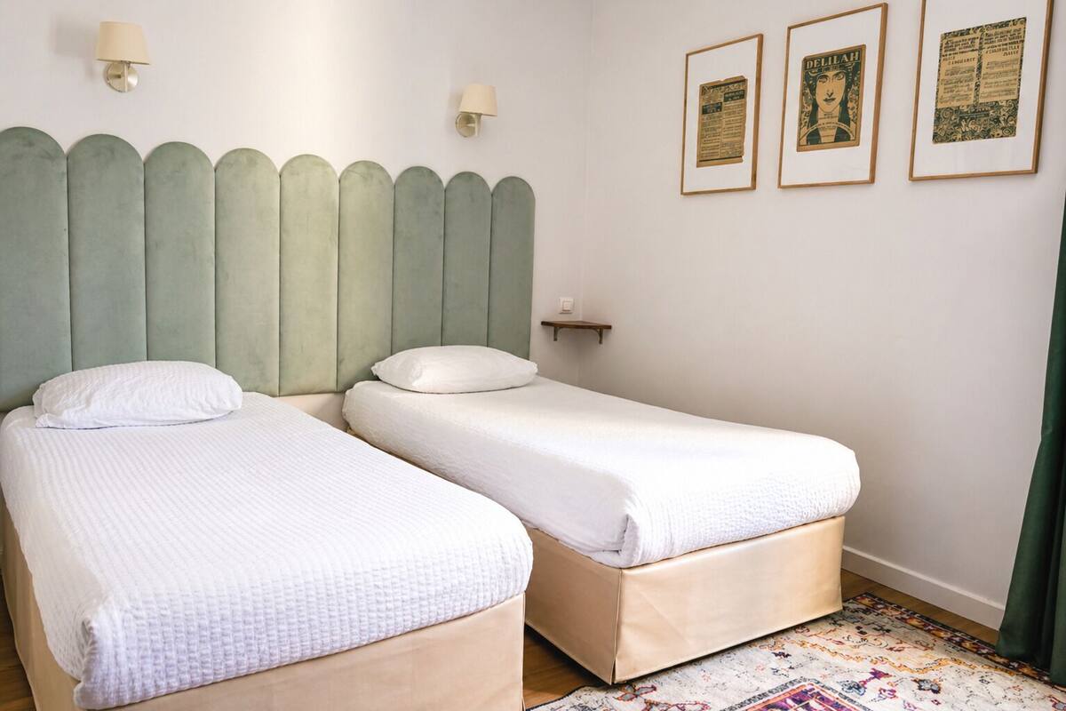 A room with two beds, a headboard, wall-mounted lamps, and framed pictures.