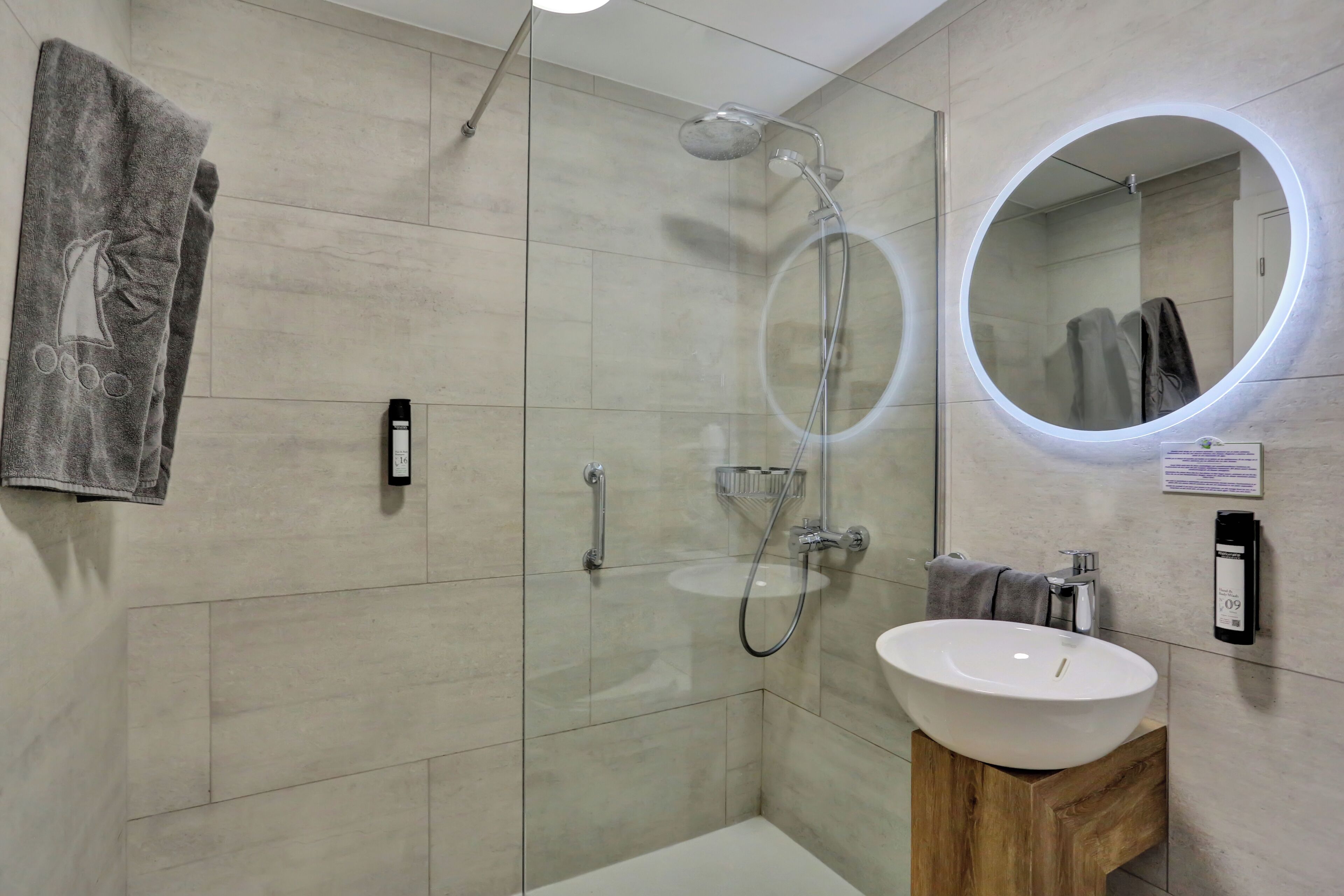 A modern bathroom with a glass-enclosed shower, a round mirror with a light, and a wooden vanity with a sink.