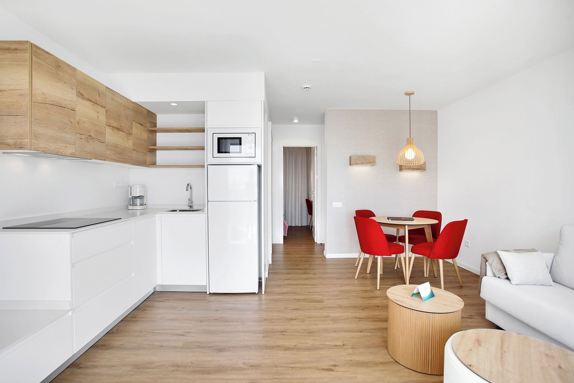 Een moderne keuken met witte kasten, een ingebouwde magnetron en een koffiezetapparaat. Er is een eethoek met een ronde tafel en rode stoelen, en een witte bank in de woonkamer.