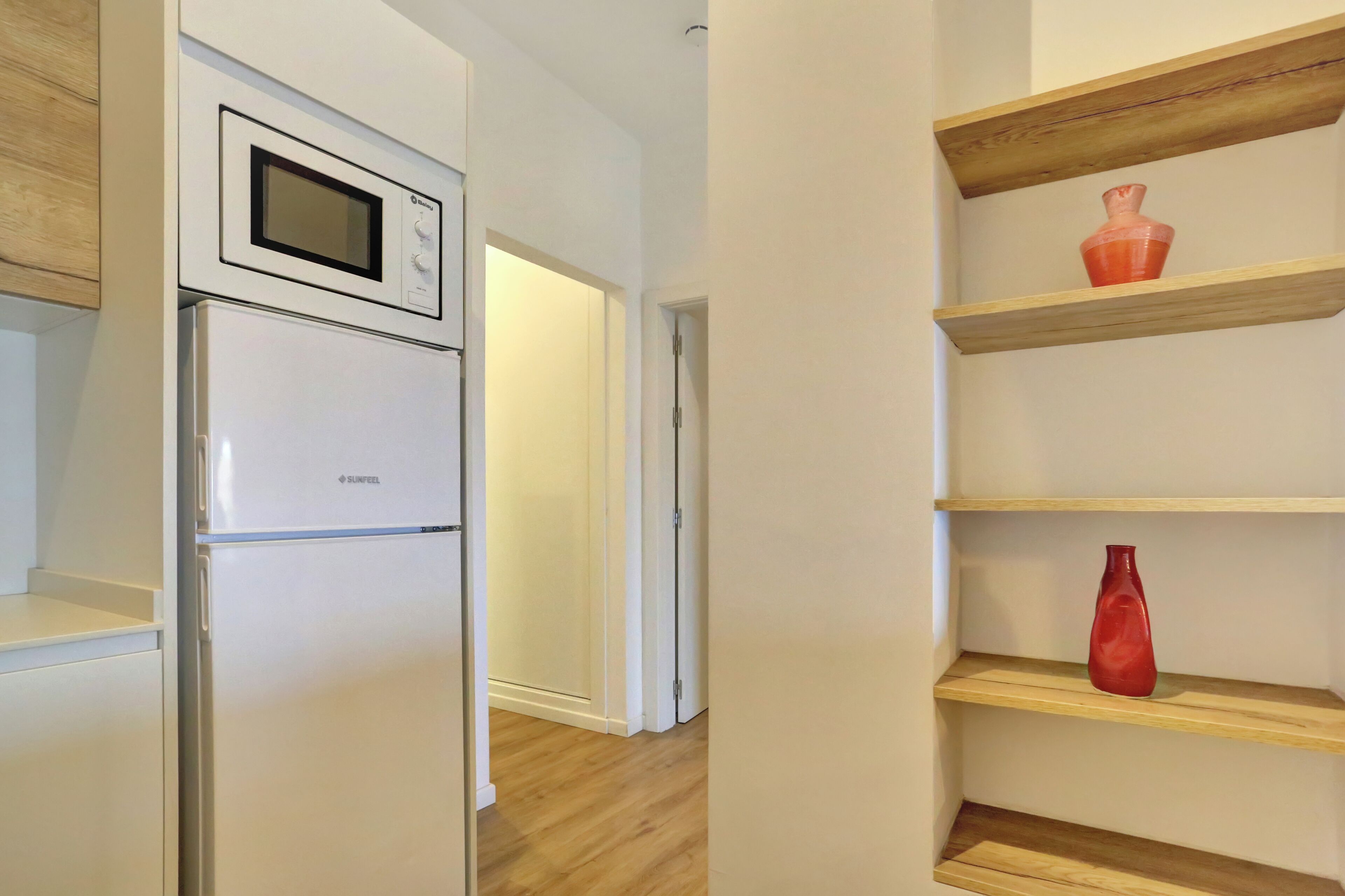 A kitchen with a built-in microwave and refrigerator, wooden flooring, and a hallway leading to another room.