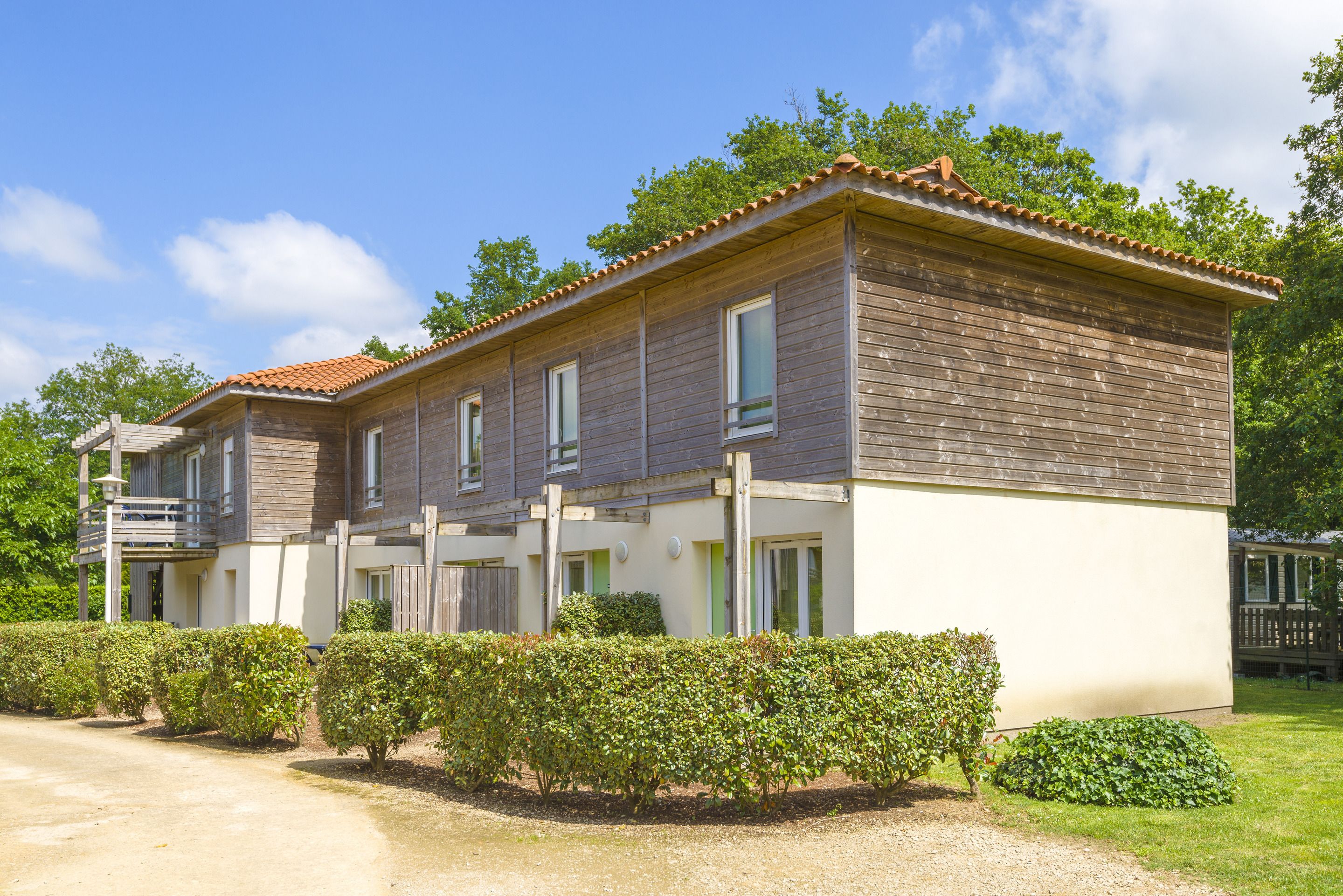 Un bâtiment à deux étages, doté d’un balcon sur le côté gauche, d’un toit recouvert de tuiles et d’un jardin bien entretenu avec des haies.