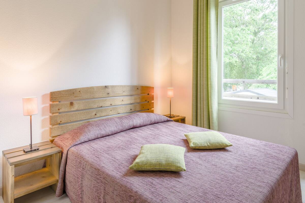 A bedroom with a wooden headboard, a bed with a pink bedspread, two pillows, a nightstand with a lamp, and a window with a view of trees.