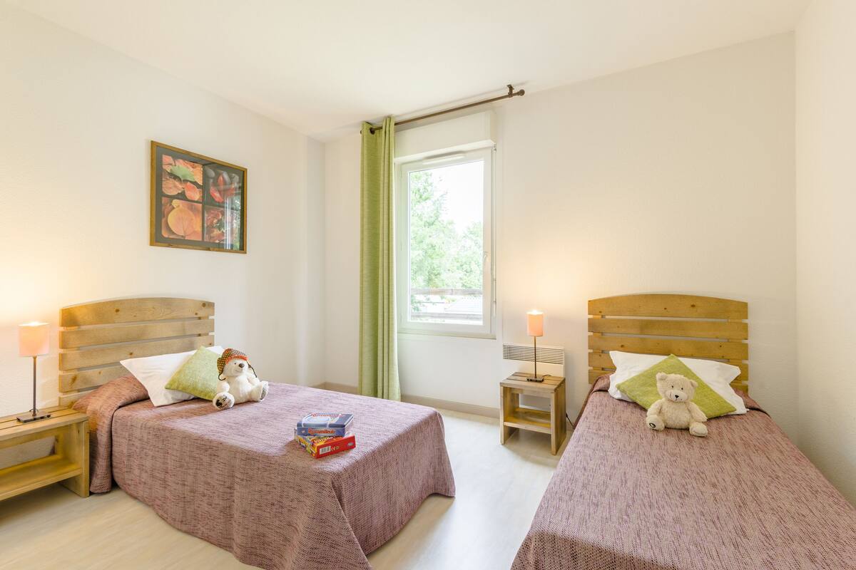 A bedroom with two single beds, each with a wooden headboard, a bedside table, a lamp, and a window with a curtain.