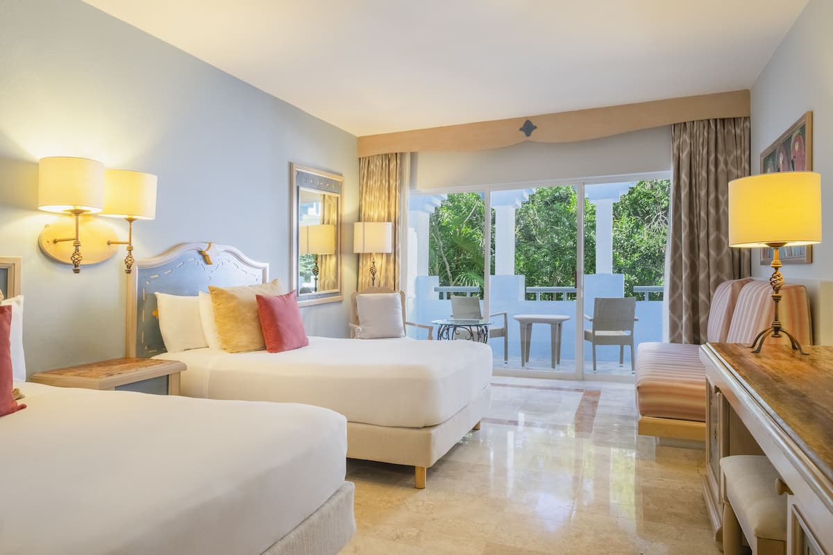 A hotel room with two beds, a desk, and a balcony with a view of greenery.