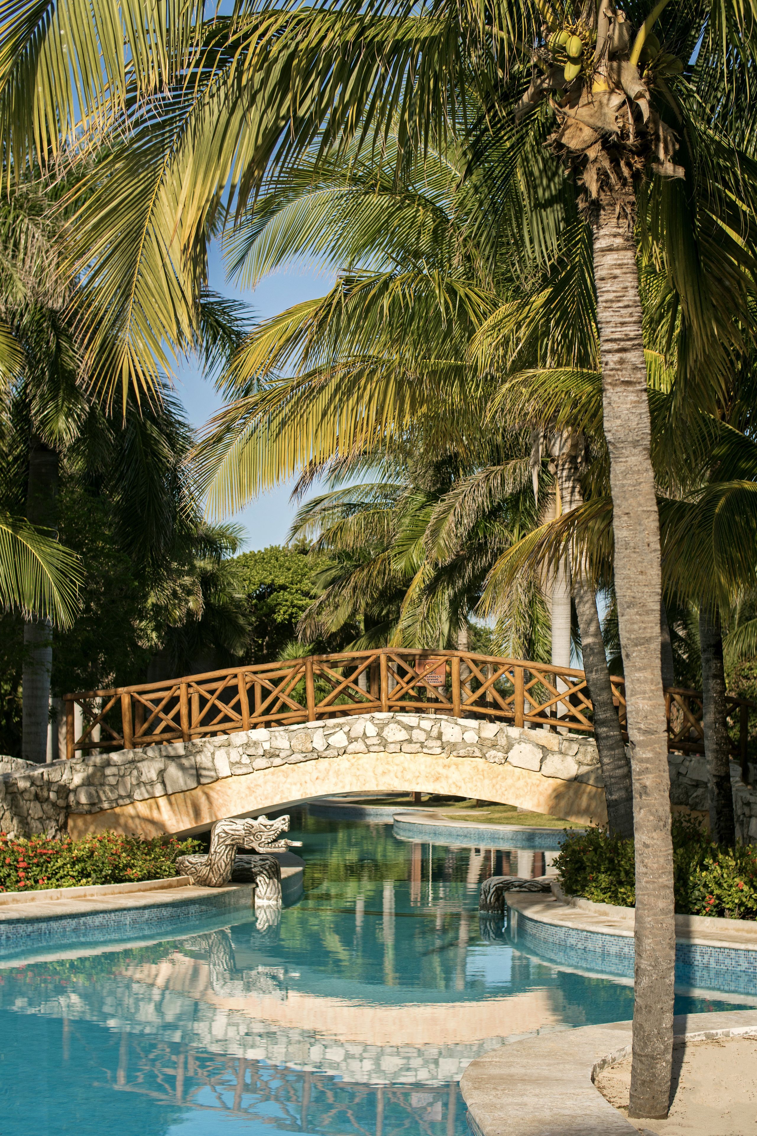 A pool with a stone bridge, palm trees, and a decorative stone sculpture.