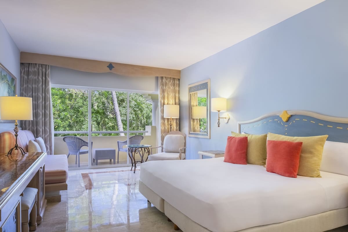 A hotel room with a large bed, a desk, a chair, and a view of greenery through the window.