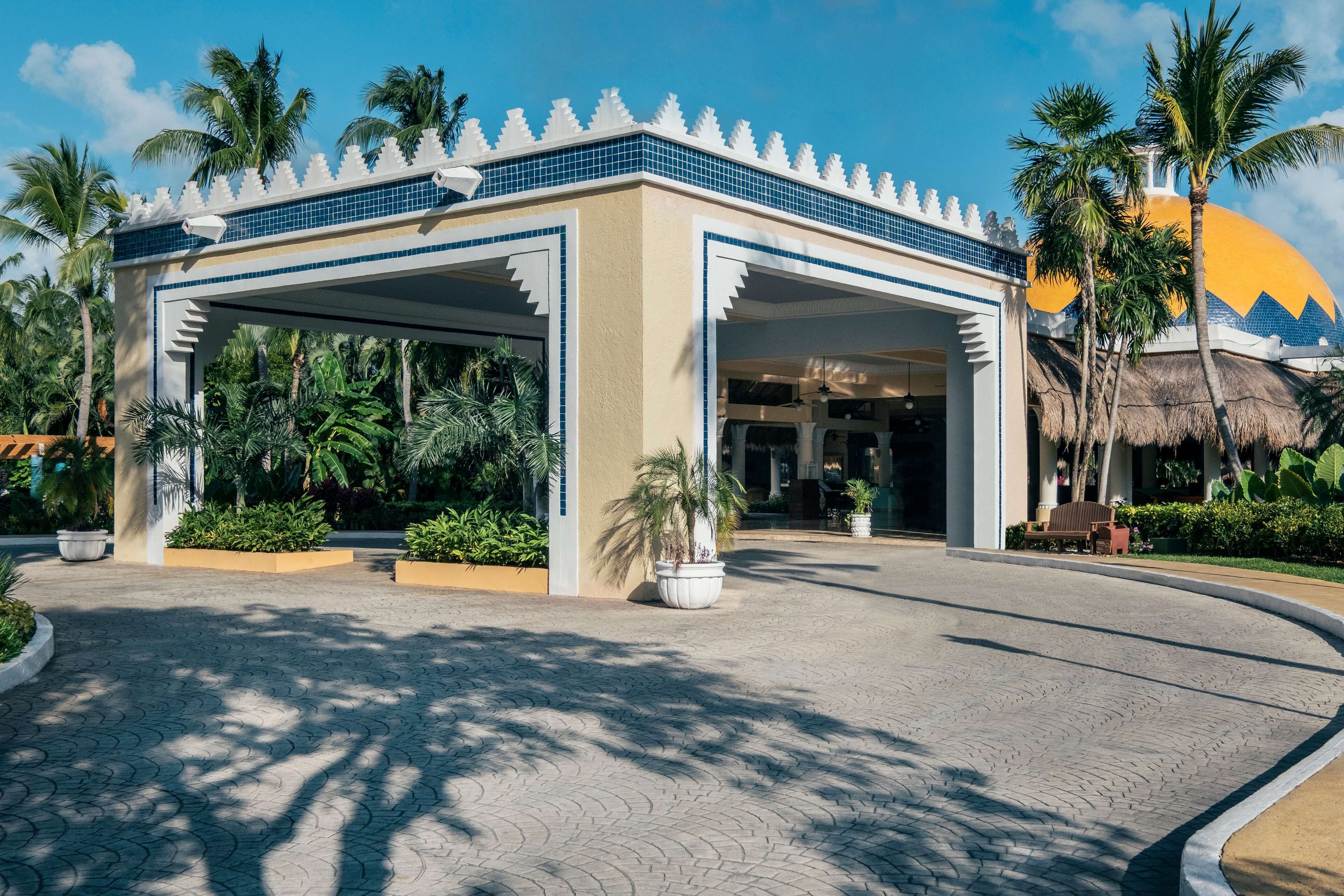 A hotel entrance with a covered area, palm trees, and a thatched-roof structure in the background.