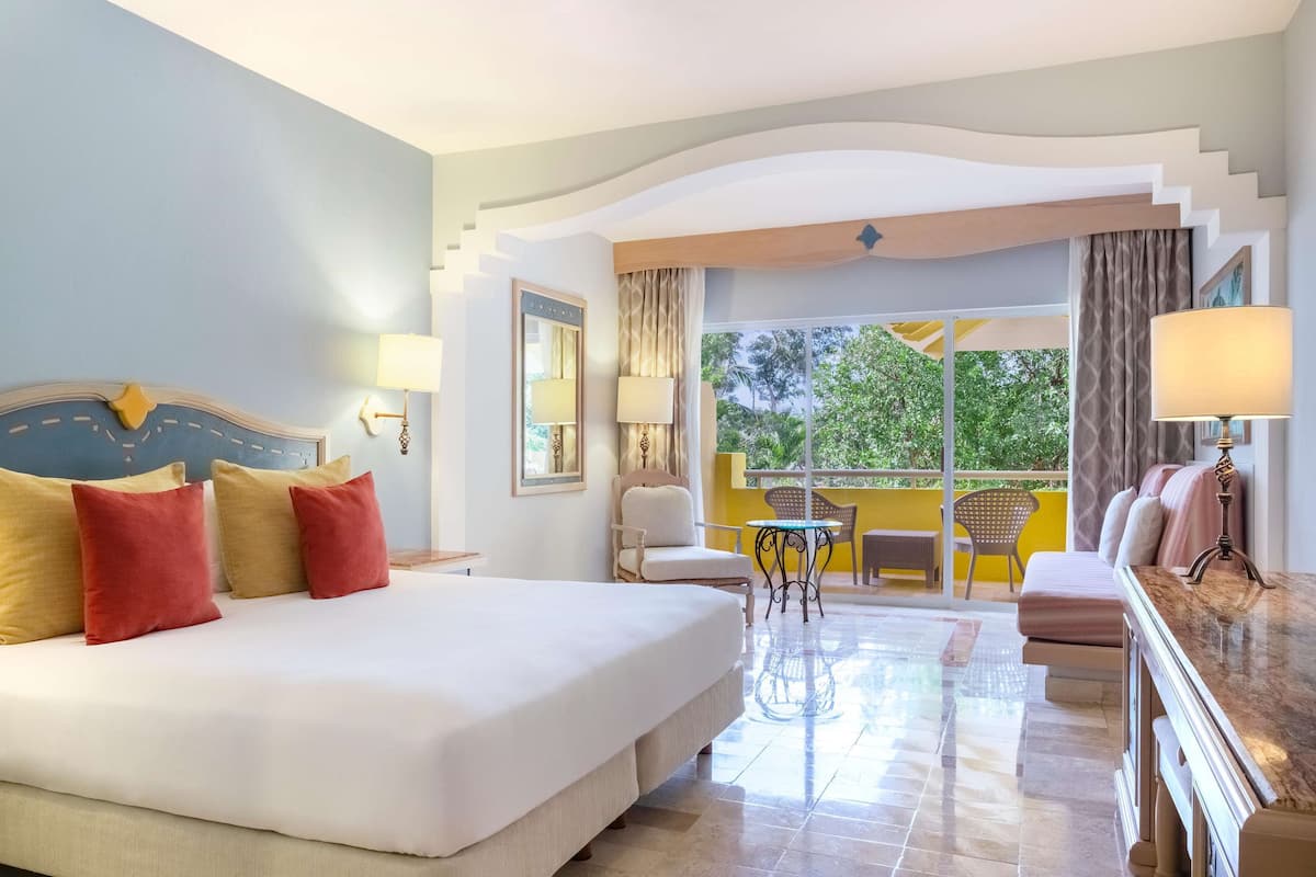 A hotel room with a large bed, a sitting area with a table and chairs, and a balcony with a view of greenery.
