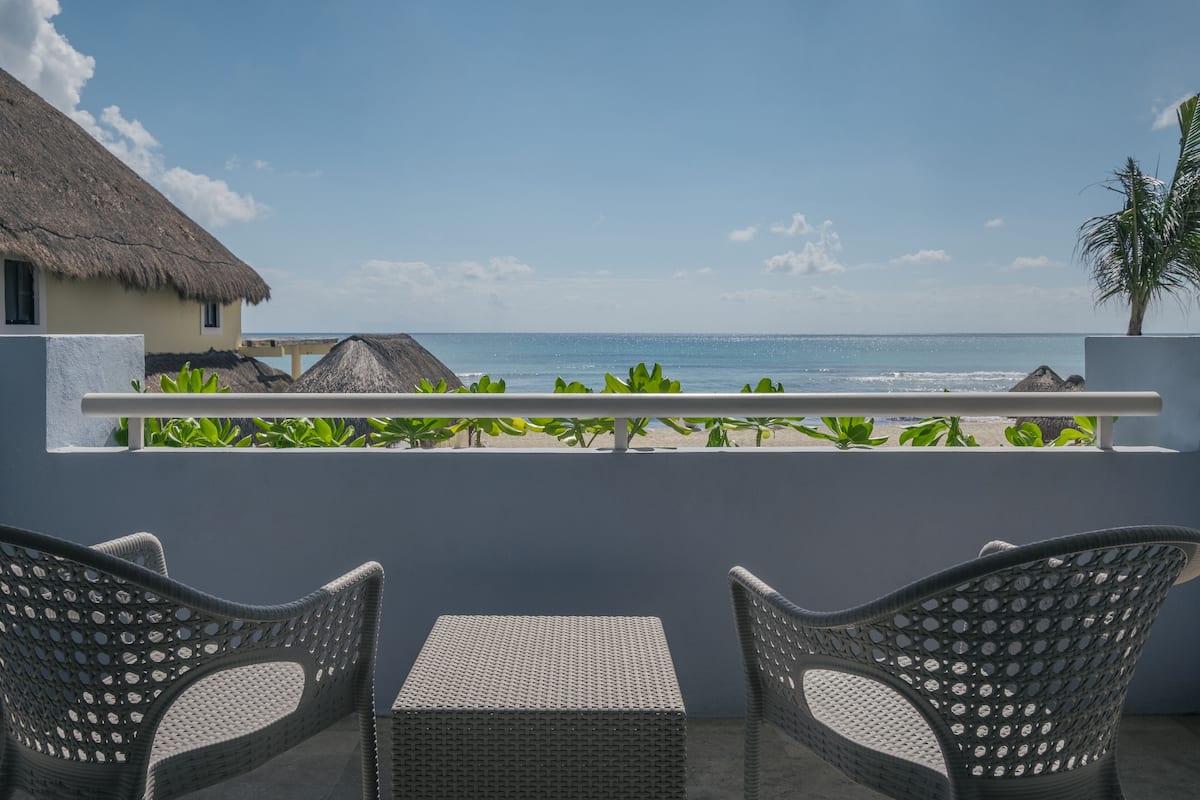 A balcony with wicker chairs and a table overlooking a beach with thatched-roof structures and greenery.