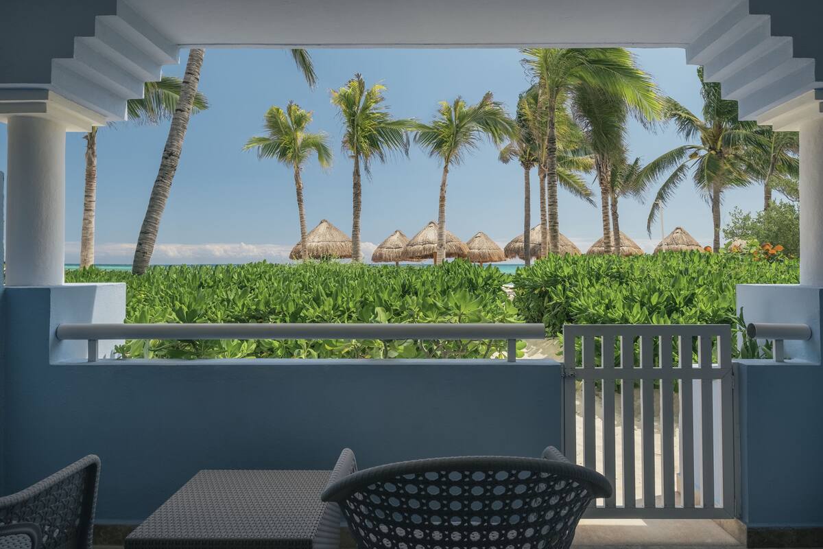 A balcony with a view of a beach, palm trees, and thatched umbrellas.