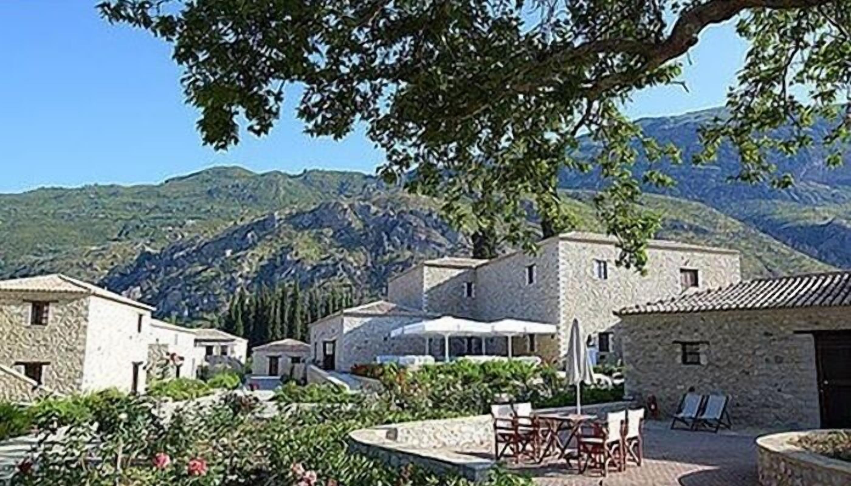 A stone building with a tiled roof, surrounded by lush greenery and a mountainous backdrop.