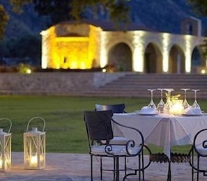 Un espacio exterior para comer con una mesa dispuesta para una comida, rodeada de sillas y faroles, con un edificio al fondo.