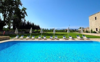 Una piscina con sillas de descanso y sombrillas, rodeada de césped y árboles, con un edificio de piedra al fondo.