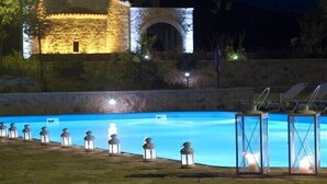 A swimming pool with lanterns and lounge chairs, a stone building with an arched entrance in the background, and a clear night sky. - Semantron Traditional Village (Aigialeia)