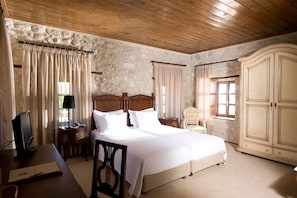 Un dormitorio con techo de madera, paredes de piedra, una cama con ropa de cama blanca, un escritorio con ordenador, una silla y un armario.