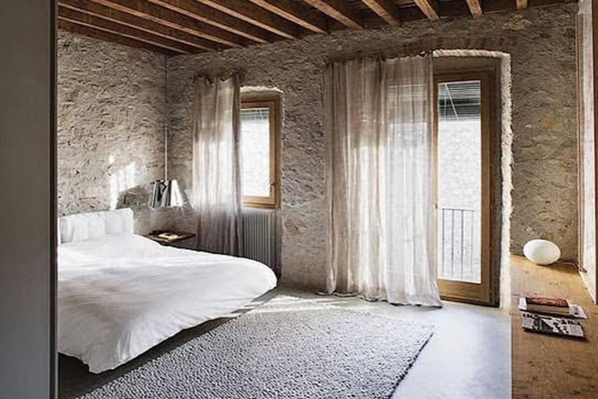 Een slaapkamer met een stenen muur, houten plafond, een bed, een raam en uitzicht op de omgeving.