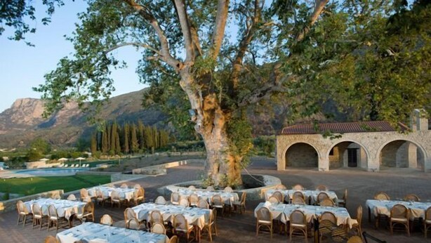 An outdoor dining area with tables and chairs, surrounded by trees and a stone building. - Semantron Traditional Village (Aigialeia)