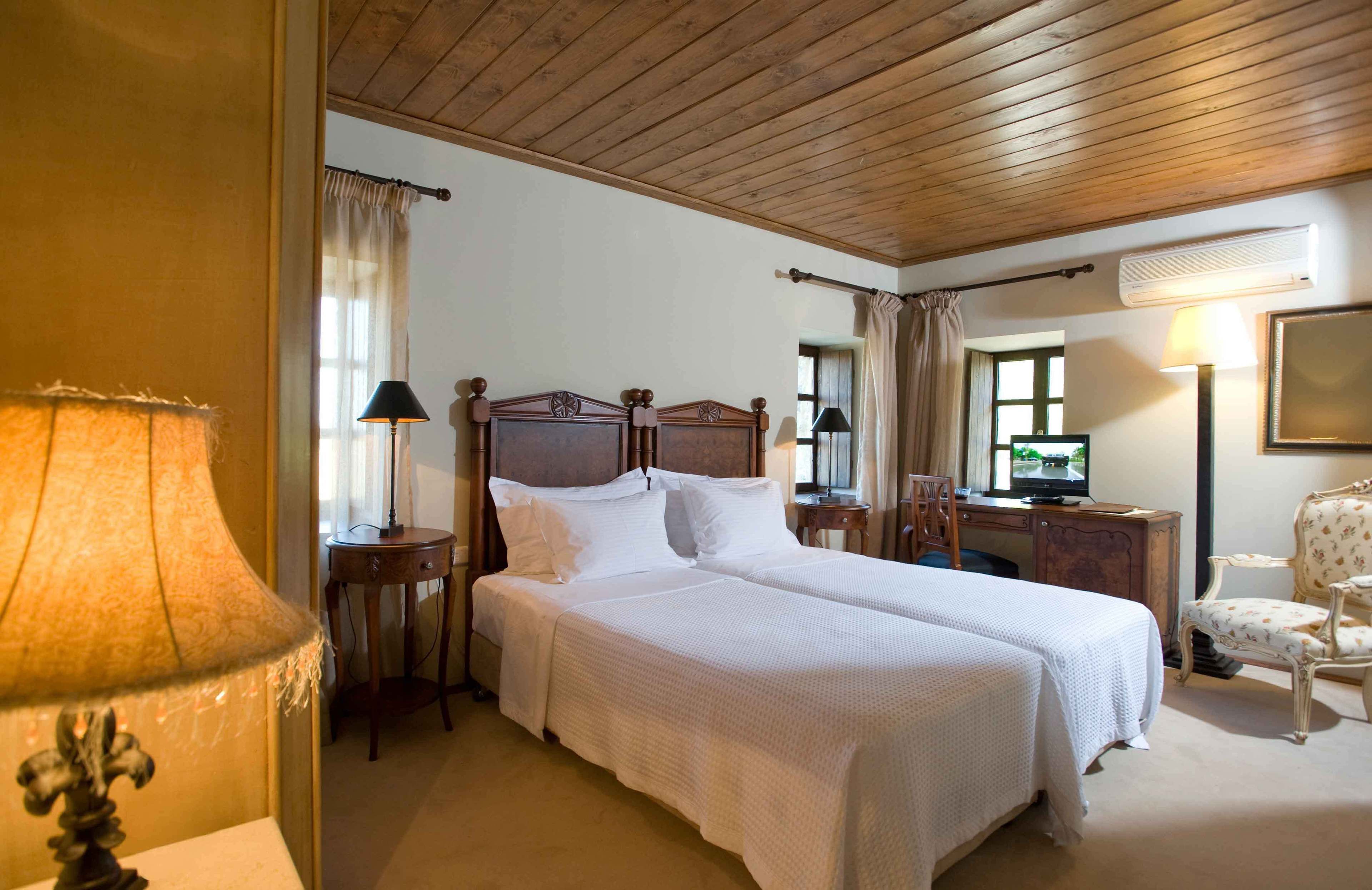 A hotel room with a wooden ceiling, a double bed with white bedding, a desk with a television, and a chair.