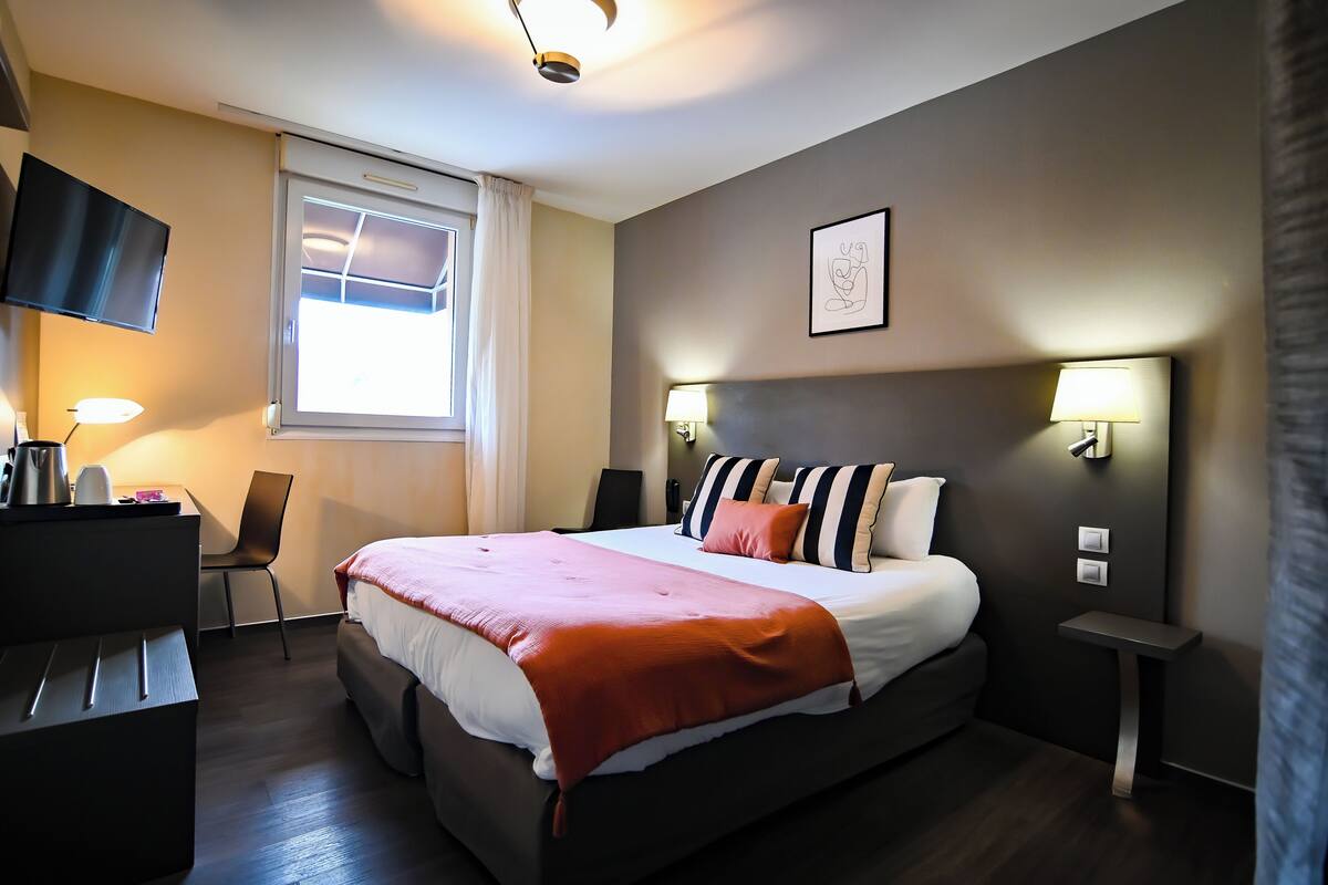 Une chambre d’hôtel comprenant un lit, des tables de chevet, un bureau, une chaise, une télévision et une fenêtre avec des rideaux.