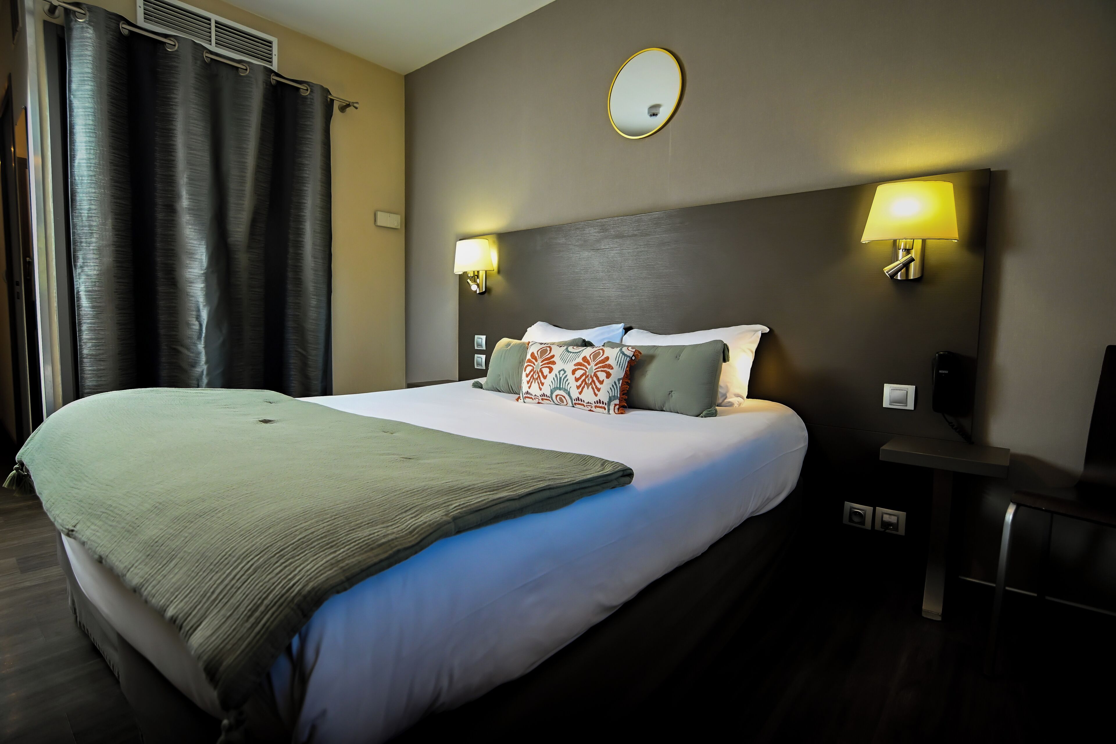 Habitación de hotel con una cama grande, dos lámparas de pared, un espejo redondo y un escritorio pequeño con una silla.