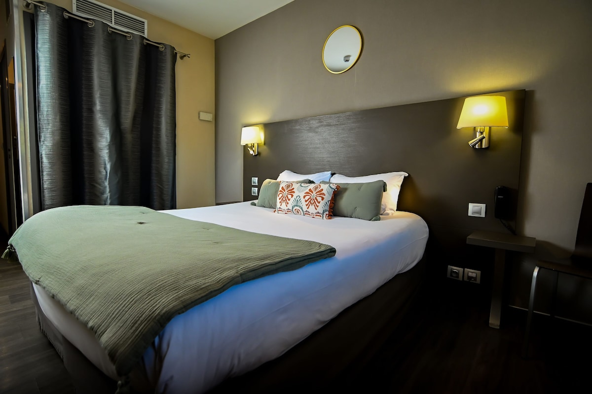 A hotel room with a large bed, two bedside lamps, a round mirror above the bed, and a small table with a chair.