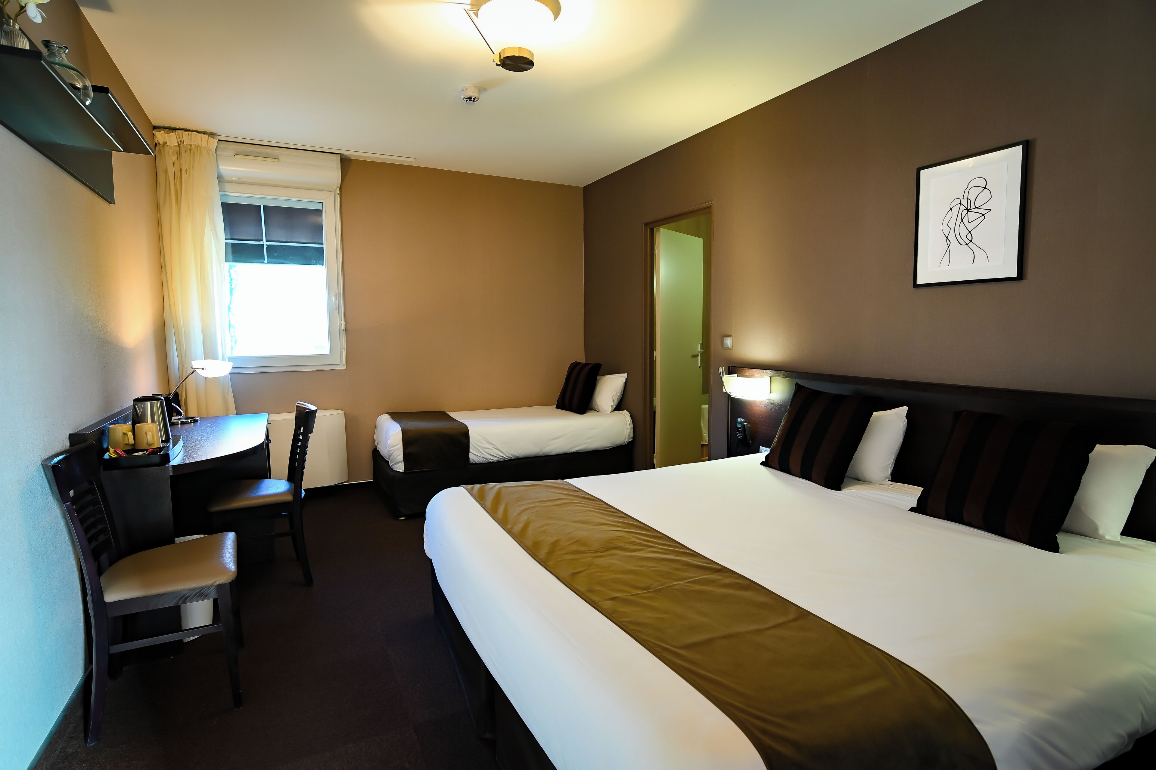 A hotel room with two beds, a desk with a chair, a window with curtains, a framed picture on the wall, and a ceiling light.