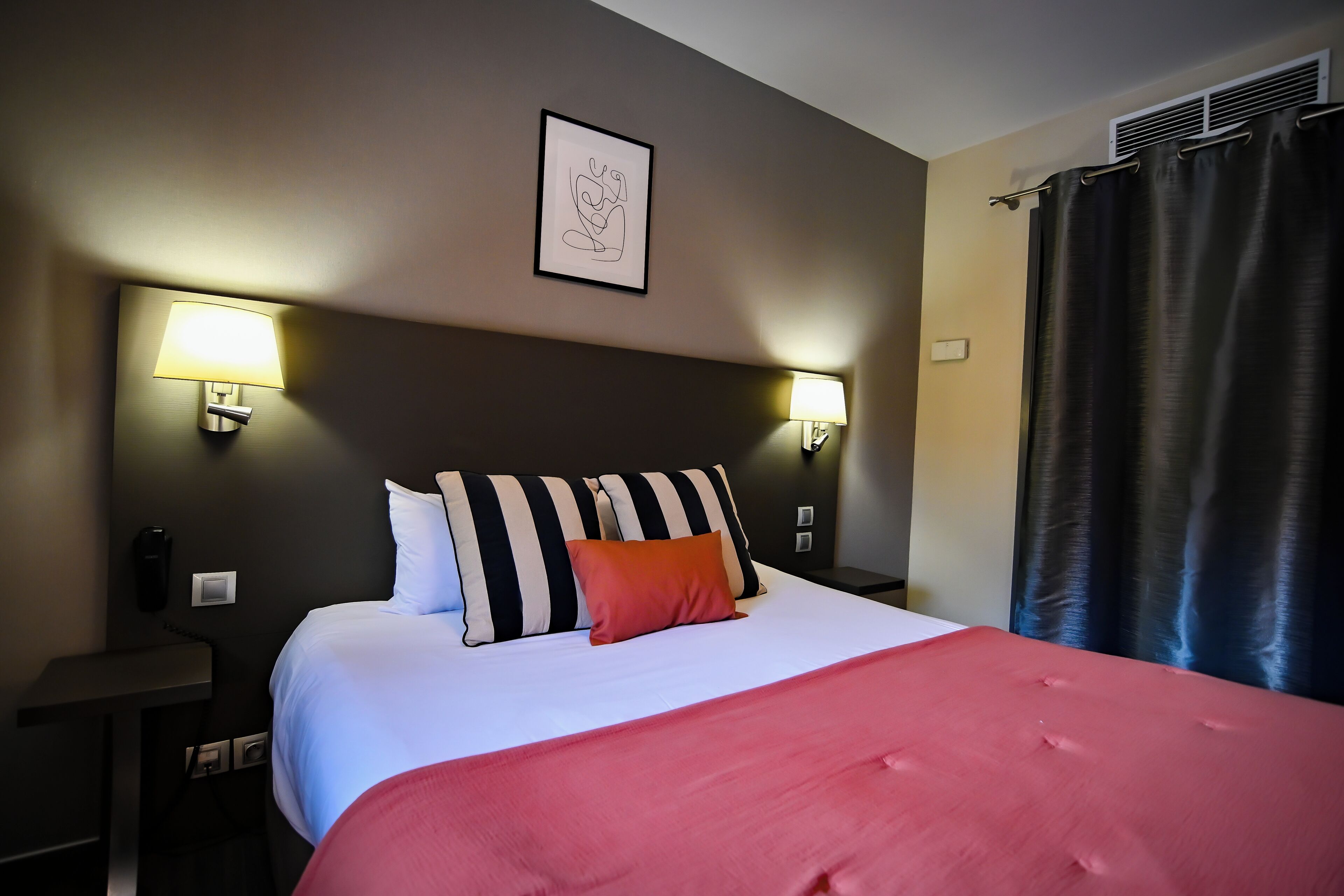 A hotel room with a bed, two bedside lamps, a wall-mounted artwork, and a window with curtains.