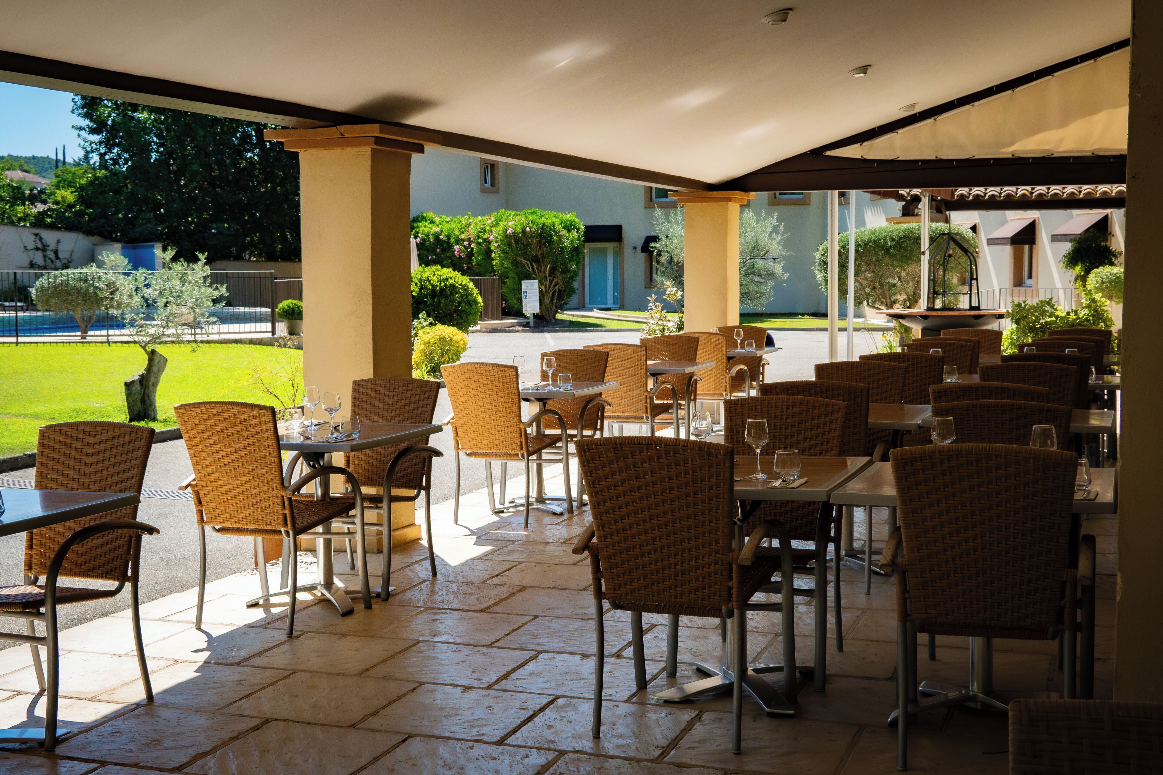 An outdoor cafe area with wicker chairs and tables, a tiled floor, and a view of a garden and building in the background.