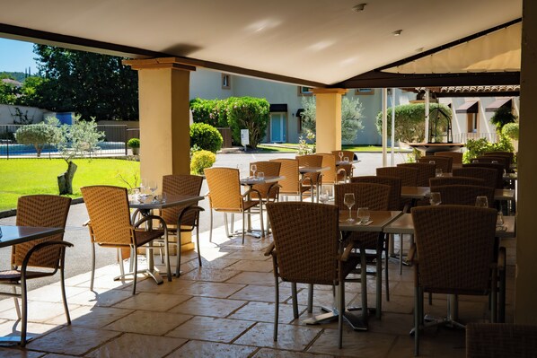 An outdoor cafe area with wicker chairs and tables, a tiled floor, and a view of a garden and building in the background. - Le Mas de Jossyl (La Roque-d'Antheron)