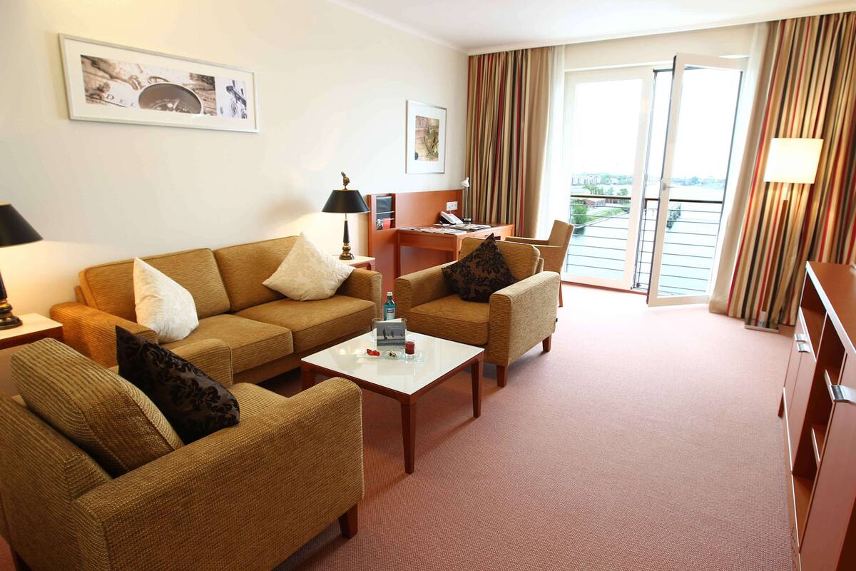 A hotel room with a sofa set, a coffee table, a desk, and a view of the city through the window.