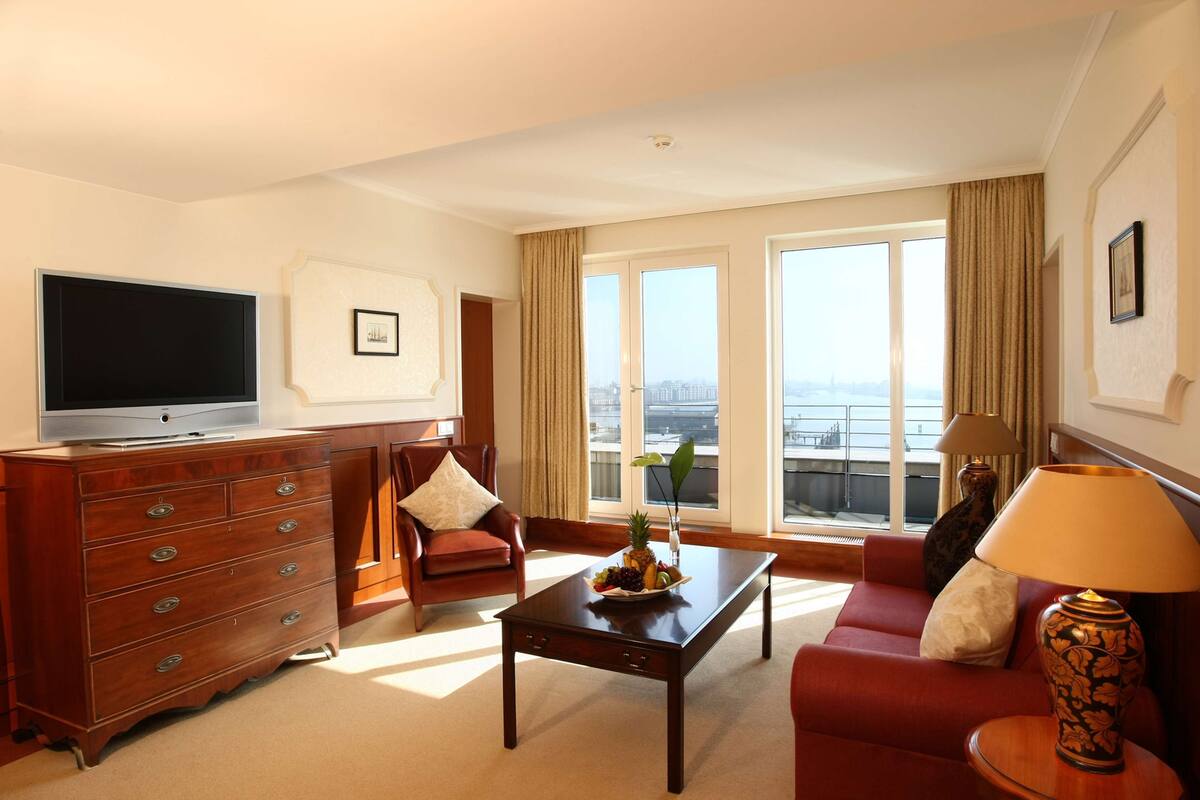 A hotel room with a television, a wooden dresser, a red sofa, a coffee table, and a large window with a view of the city.