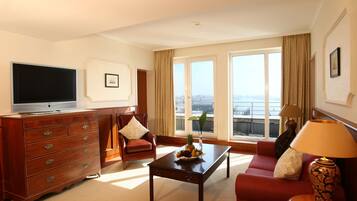 A hotel room with a television, a wooden dresser, a red sofa, a coffee table, and a large window with a view of the city.