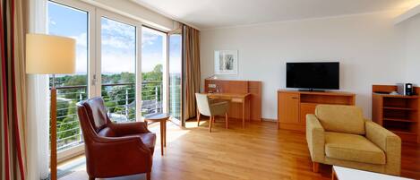 A modern hotel room with a wooden floor, a large window with curtains, a flat-screen TV, a desk, two chairs, and a bookshelf.