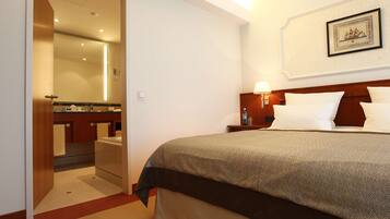 Una habitación de hotel con una cama, una mesita de noche con lámpara, un baño con espejo y un cuadro enmarcado en la pared.