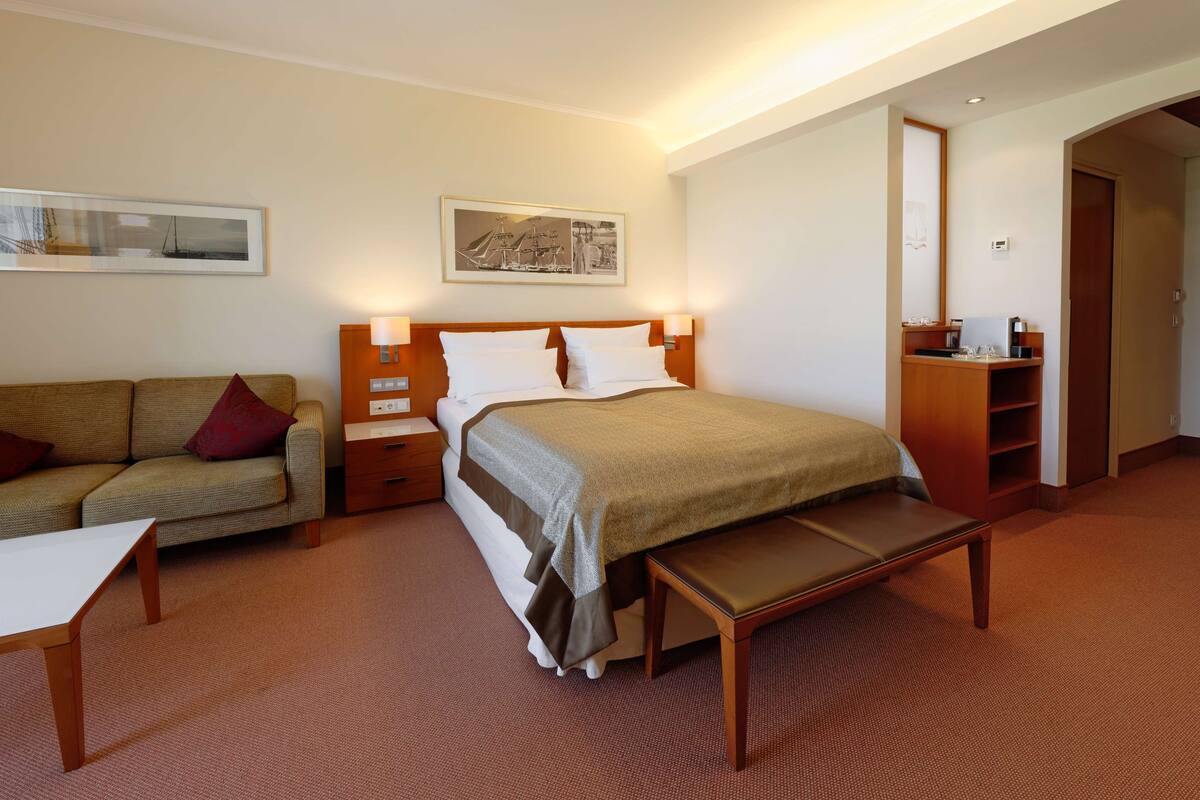 A hotel room with a bed, a sofa, a bedside table, a nightstand, a bench, a wooden cabinet, and a wall-mounted mirror.