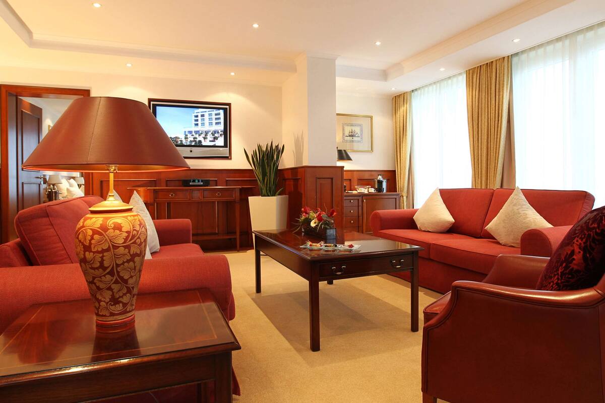 A hotel room with a red sofa, a wooden coffee table, a decorative vase, and a television on the wall.