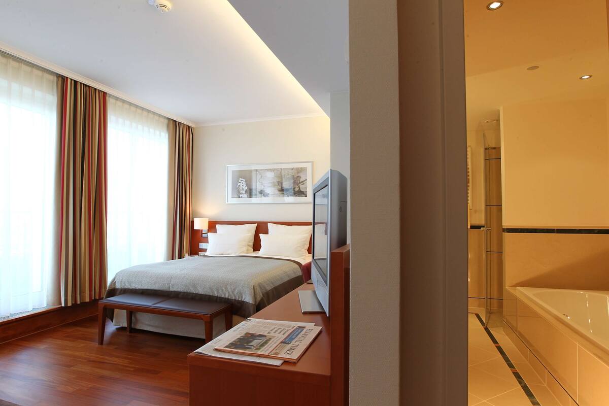 A hotel room with a bed, a bench, a television, and a bathroom with a bathtub.
