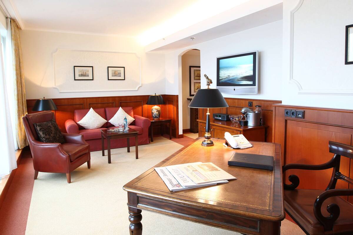A hotel room with a red sofa, a wooden coffee table, a desk with a telephone, and a television on the wall.