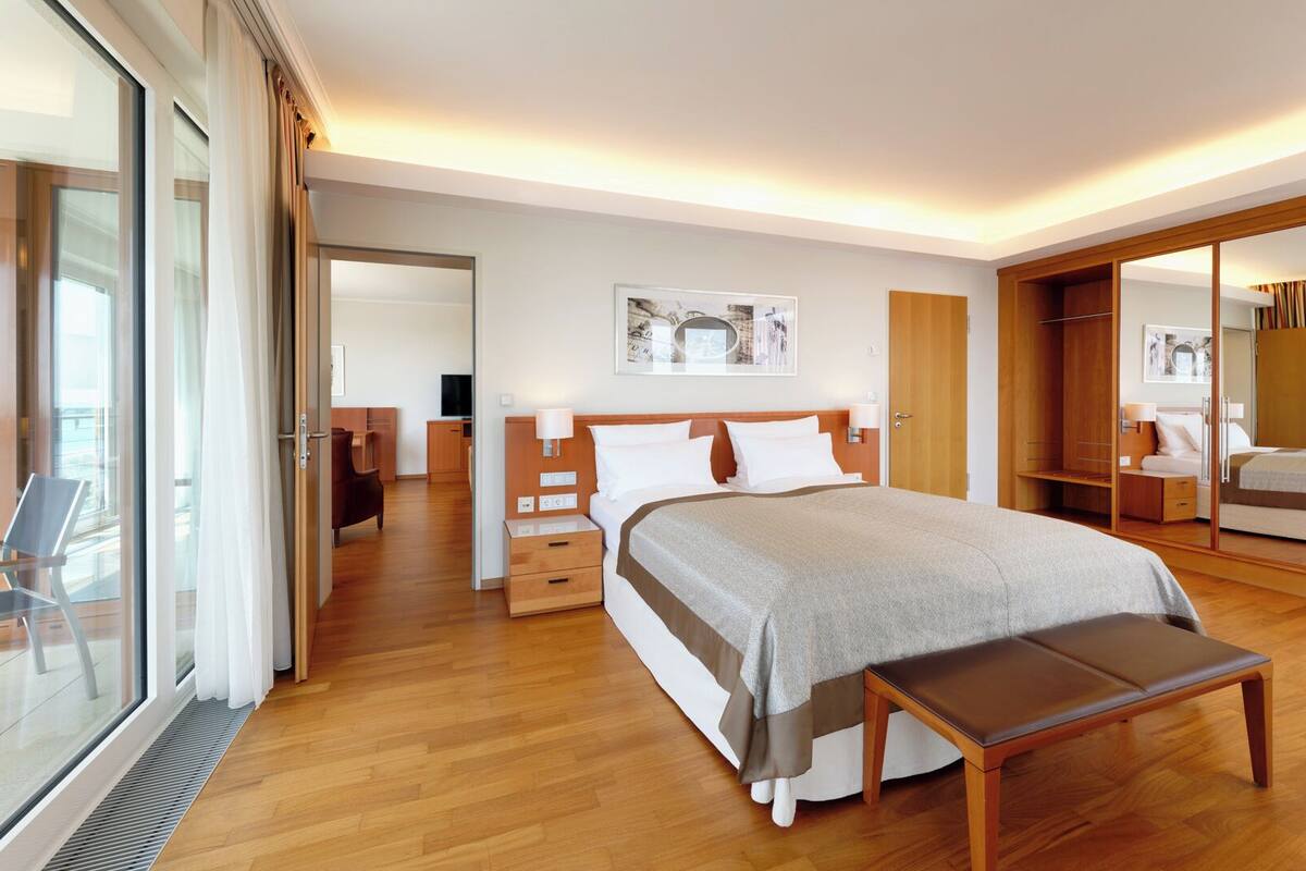 A modern hotel room with a large bed, bedside tables, a bench, a wardrobe, and a large mirror.